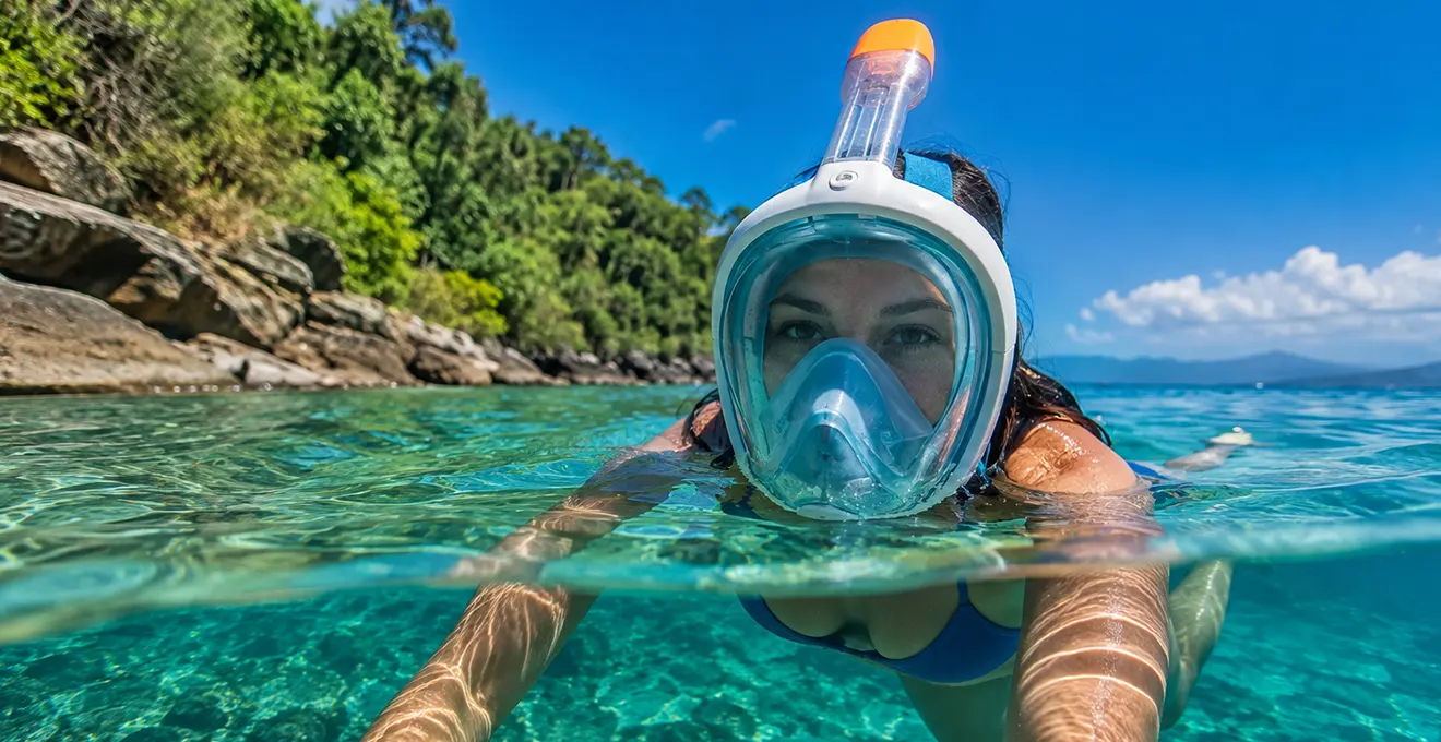 Snorkeleur équipé d'un masque facial intégral avec tuba intégré, nageant en surface dans une zone côtière peu profonde aux eaux cristallines